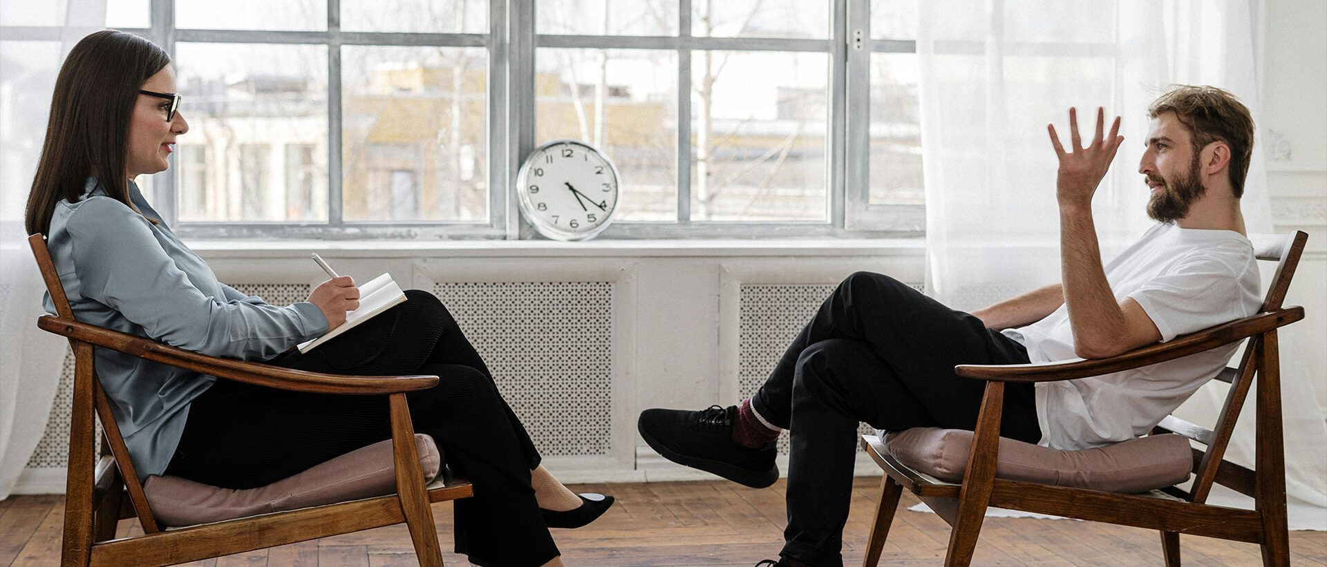 A couple sitting in chairs with one of them looking at the clock.